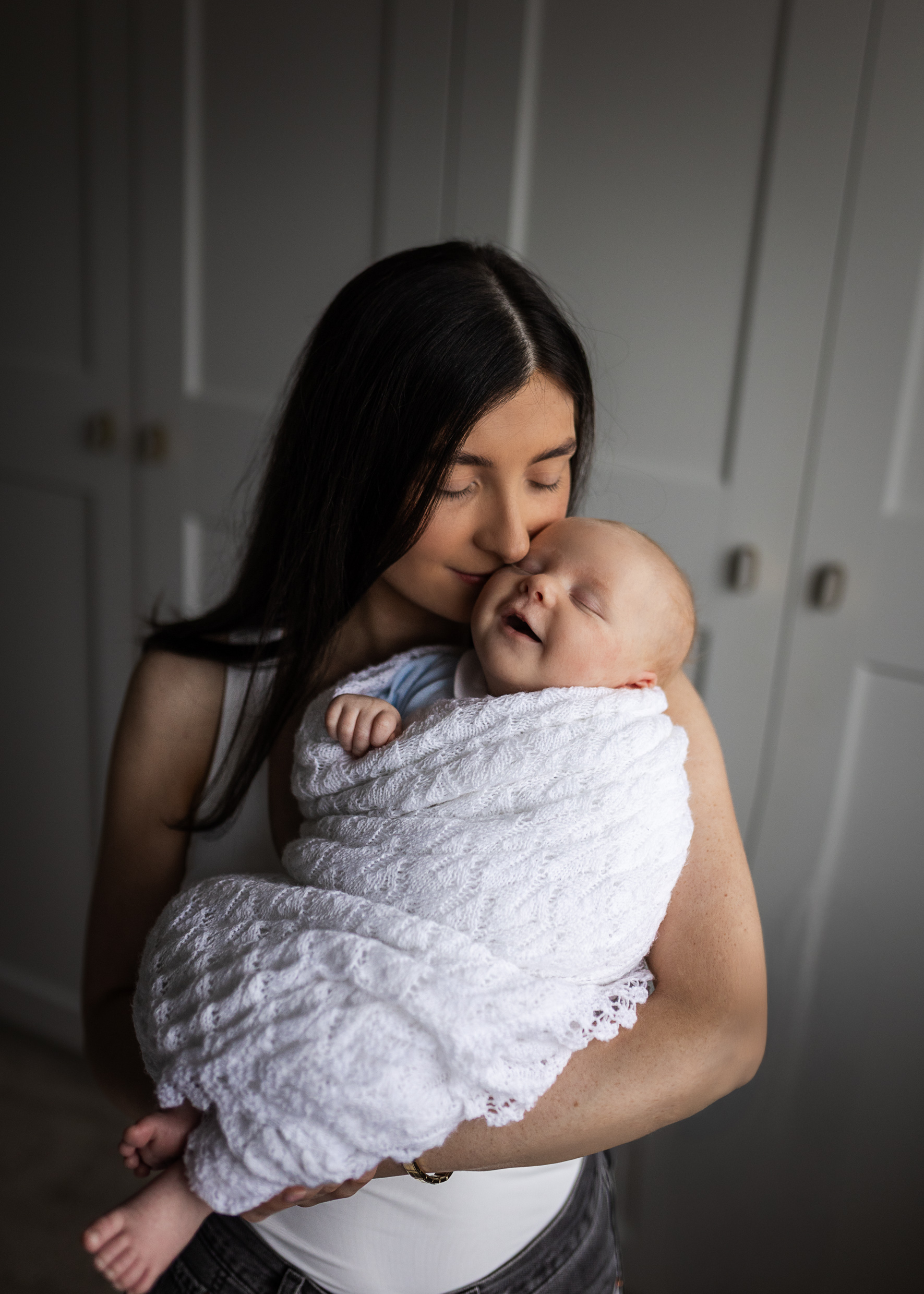 Mother cuddling wrapped newborn baby while toddler smiles beside her during in-home family session