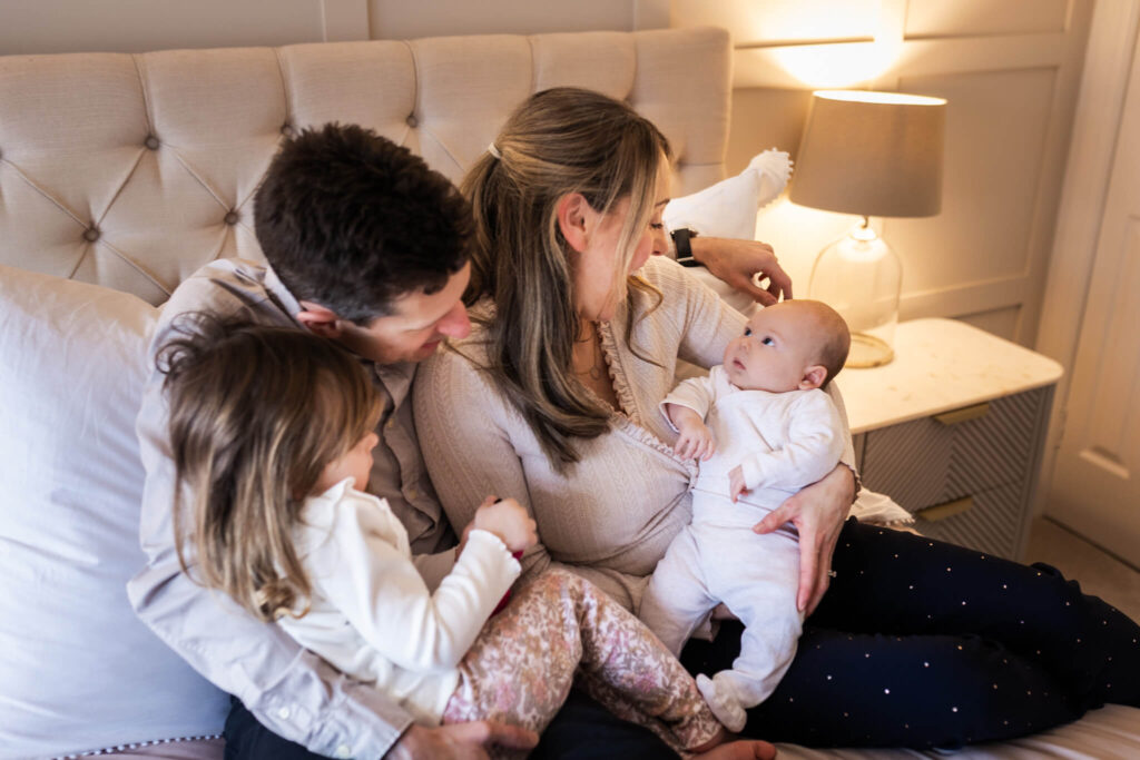 Parents sitting on a bed with their older daughter as the mother holds their newborn baby during a lifestyle newborn photoshoot in Glasgow