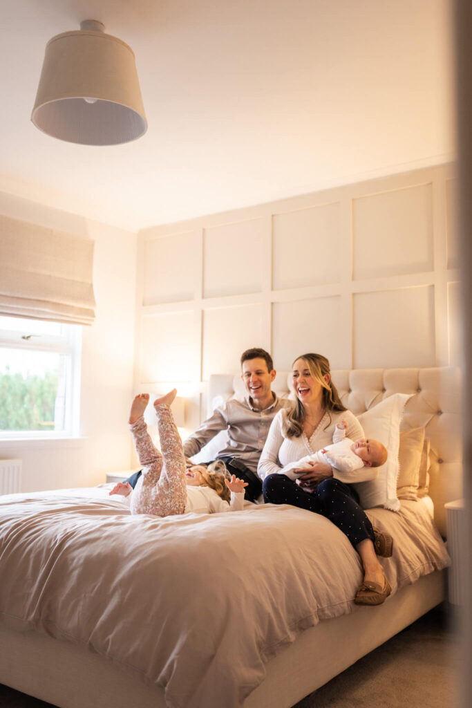 Family laughing together on bed while older sister jumps beside parents and newborn baby