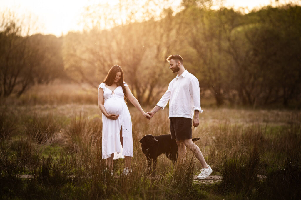 Expectant parents holding hands in Mugdock Country Park while their dog stands between them during an outdoor maternity session.