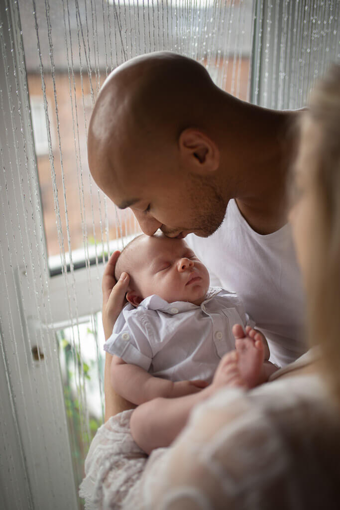 Father kissing his sleeping newborn baby's head beside a window during an at-home newborn photoshoot in Glasgow