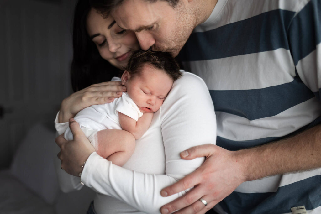 Parents cuddling their sleeping newborn baby, with dad gently kissing the baby's head during an at-home newborn photoshoot in Glasgow