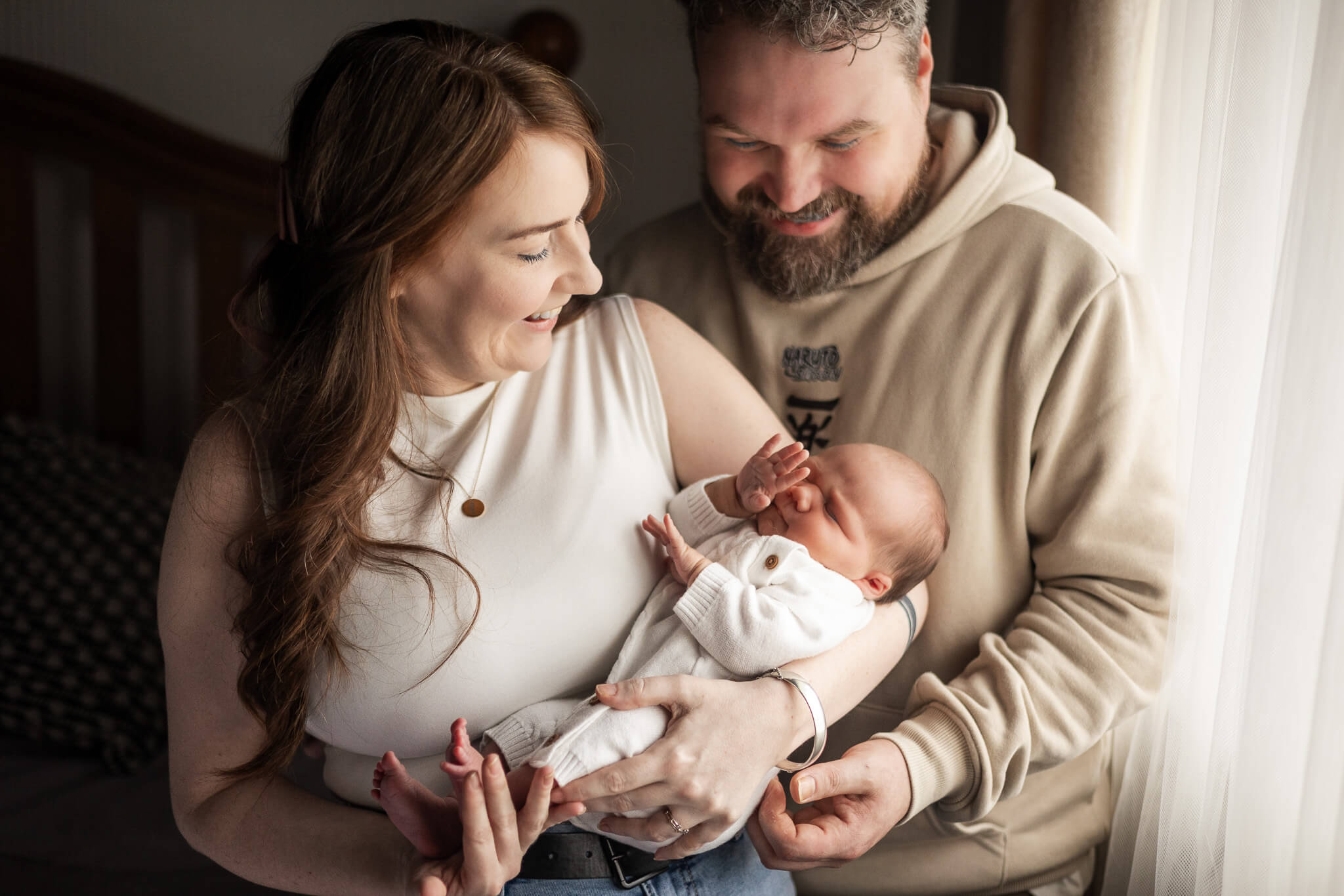 Parents looking down at their newborn baby together beside a window during an at-home newborn photoshoot in Glasgow