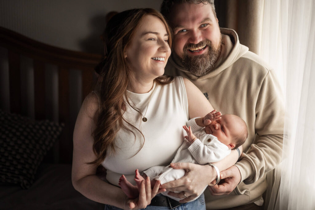 Smiling parents holding their newborn baby together in soft window light