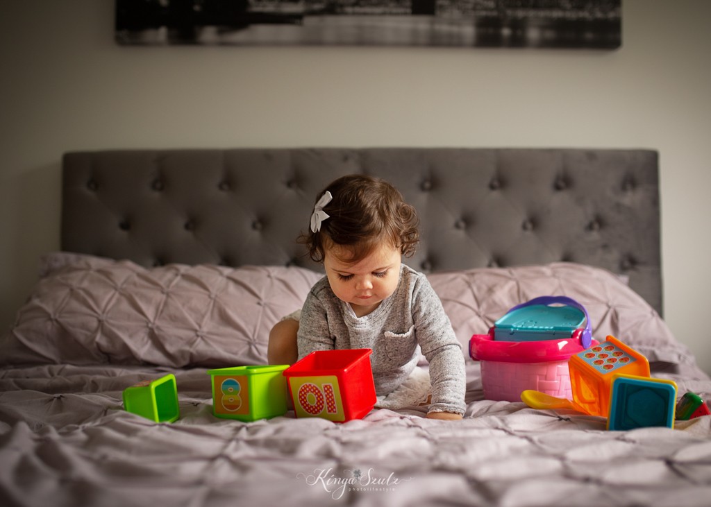 one year old baby girl sitting in the middle on the master bed playing with toys, family lifestyle photoshoot in the home