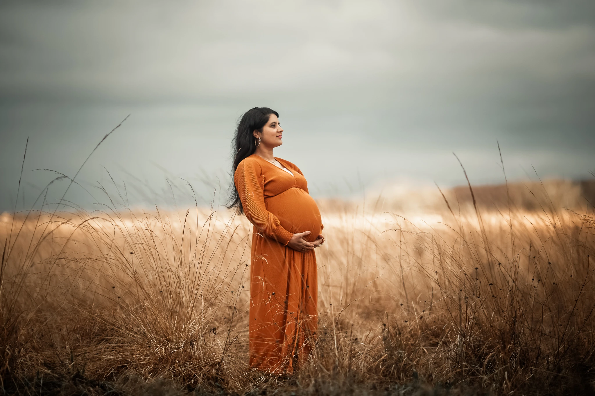Pregnant woman in an orange maternity dress holding her bump in a meadow during an outdoor maternity photoshoot in Glasgow