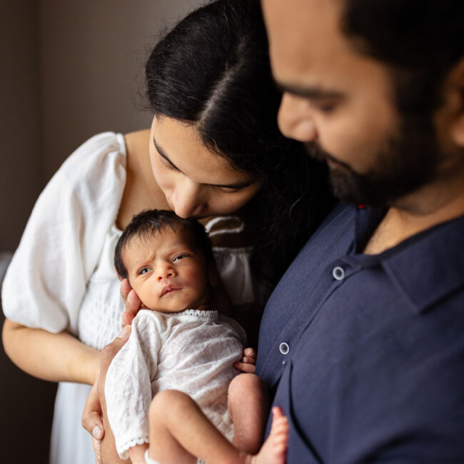 Parents cuddling their awake newborn baby, with mum leaning in to kiss the baby's head during an at-home newborn photoshoot in Glasgow