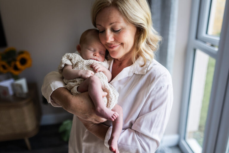 Grandmother holding a newborn baby in her arms beside a window during an at-home newborn photoshoot in Glasgow