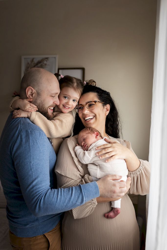 Mum holding her newborn baby while dad cuddles their older daughter, the whole family smiling together