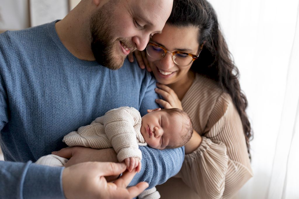 Mum leaning against dad's shoulder as he cradles their newborn baby and gently touches its tiny fingers