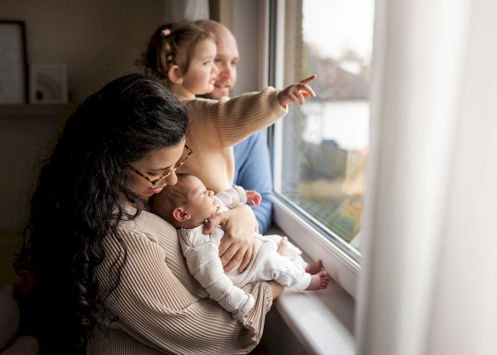 Mother holding her newborn baby by the window as her older daughter points outside during an at-home family photoshoot in Glasgow