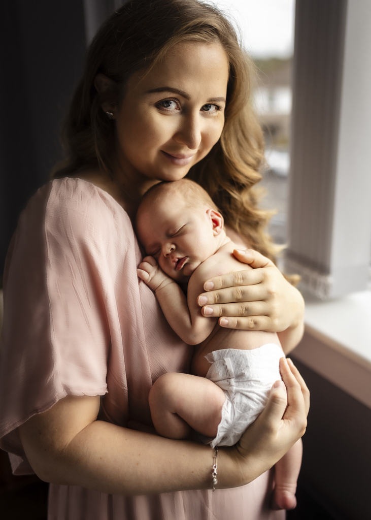 Mother holding her sleeping newborn baby beside a window during an at-home newborn photoshoot in Glasgow