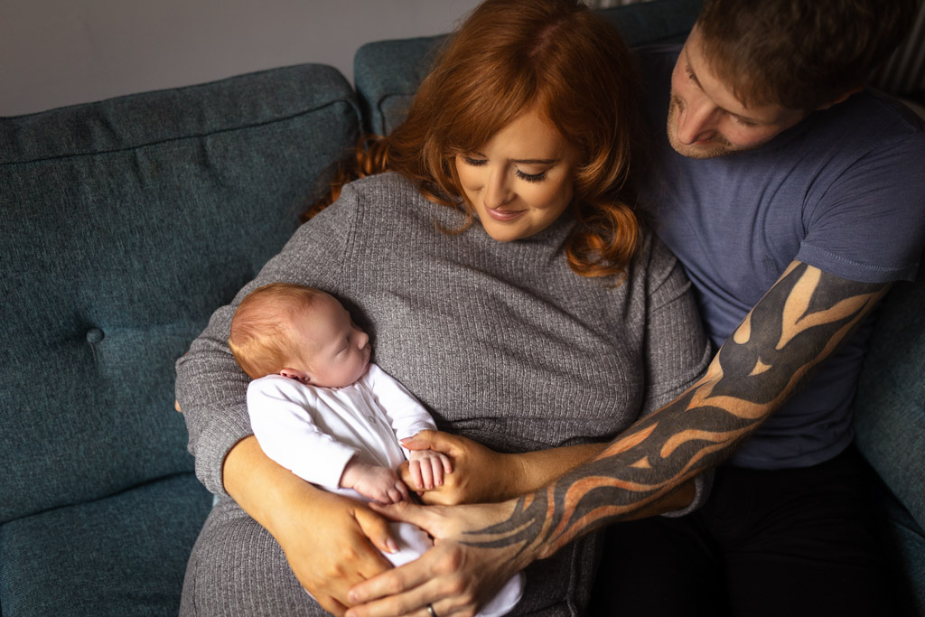 Mum holding her newborn baby on the sofa while dad wraps his arms around them during a relaxed home newborn session