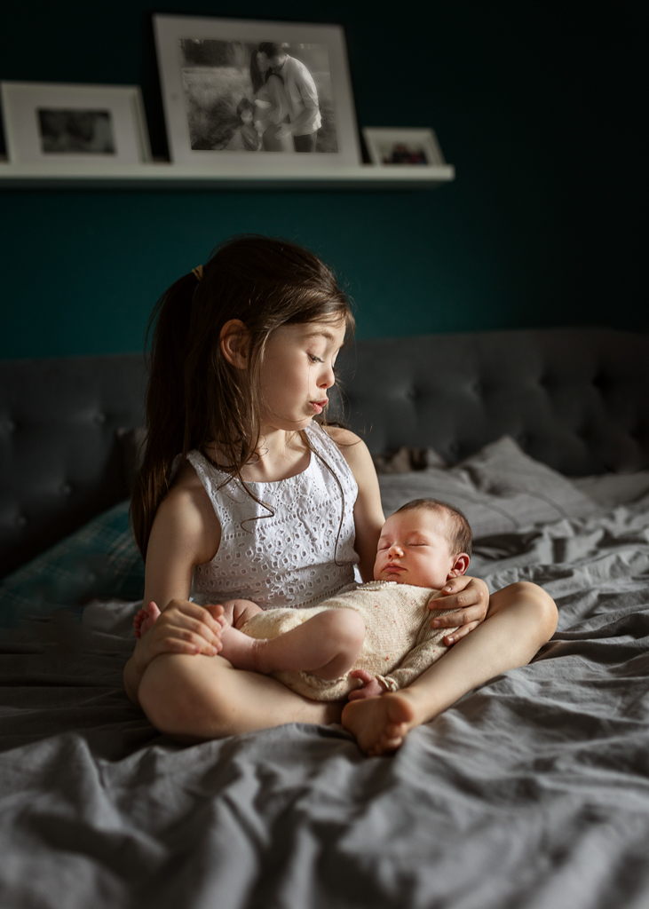Older sister holding her sleeping newborn sibling while sitting on the bed during a family newborn photoshoot in Glasgow