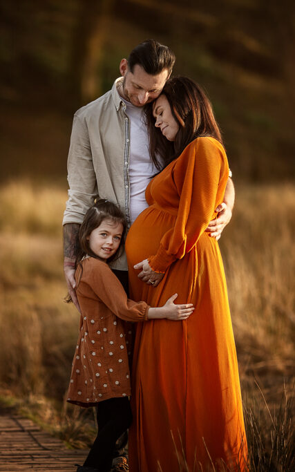 Pregnant mum in an orange dress hugging her young daughter in golden grass during a family maternity photoshoot in Glasgow