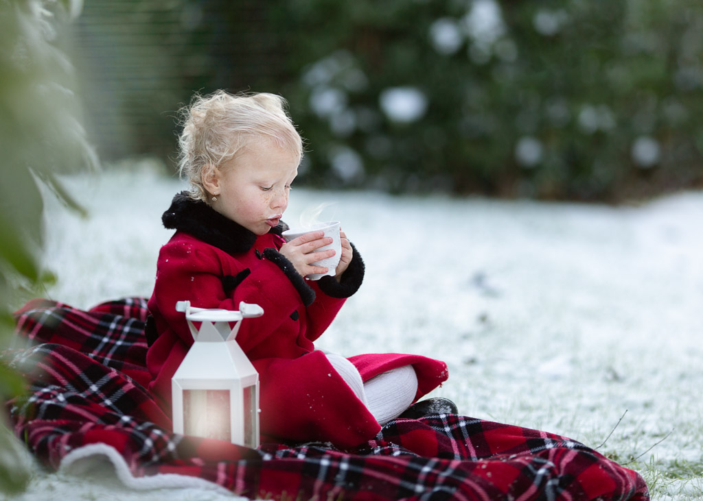 winter portrait, a 4-year-old girl sitting on the blanket in the snow drinking warm milk form the cup.