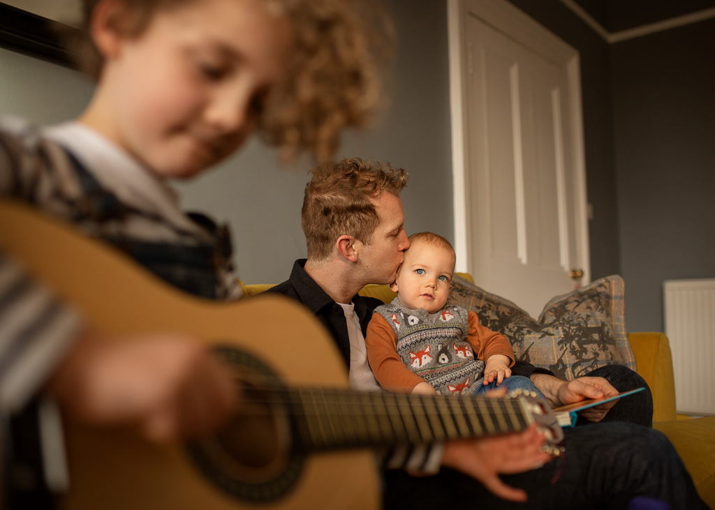 family of three, dad with son sit and listen to daughter playing guitar