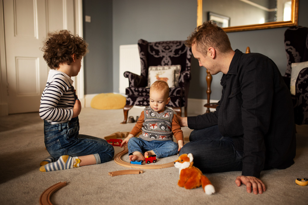 dad with son sit on the carpet and play with wooden train set during an at-home family photoshoot