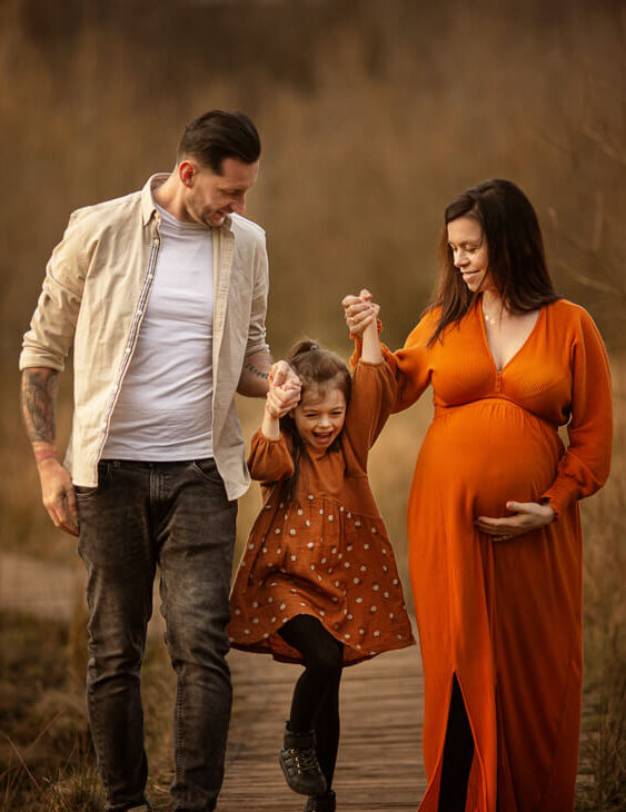 Parents holding their daughter’s hands as she jumps between them during an autumn family maternity photoshoot in Glasgow at golden hour