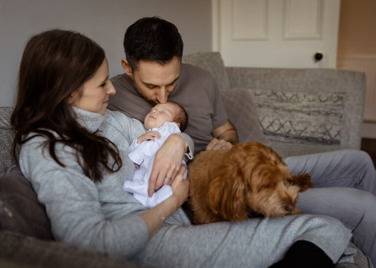 Parents cuddling their newborn baby on the sofa as dad kisses the baby's head, with their dog beside them during an at-home newborn photoshoot in Glasgow