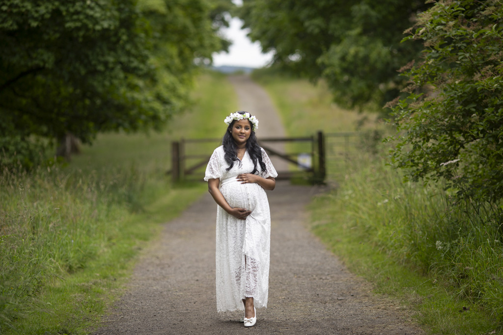Pregnant woman in a white lace dress and flower crown holding her bump on a woodland path during a maternity photoshoot in Glasgow