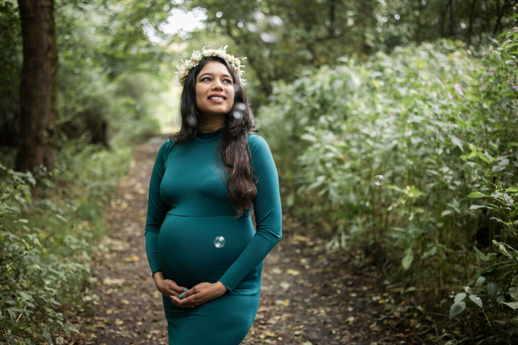 Pregnant woman in a green maternity dress and flower crown standing on a woodland path during an outdoor maternity photoshoot in Glasgow
