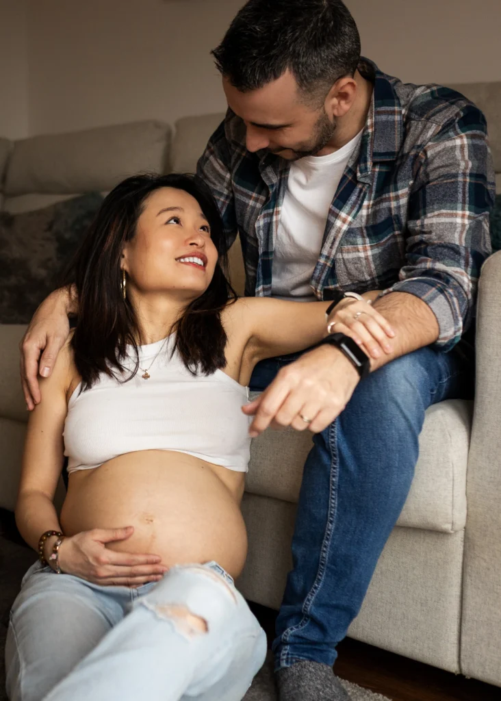 Pregnant woman sitting on the sofa with her partner as he gently holds her bump during an in-home maternity photoshoot in Glasgow