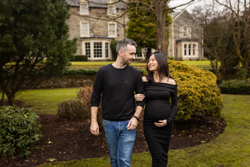 Pregnant woman in a black dress walking arm in arm with her partner in a garden during an outdoor maternity photoshoot in Glasgow