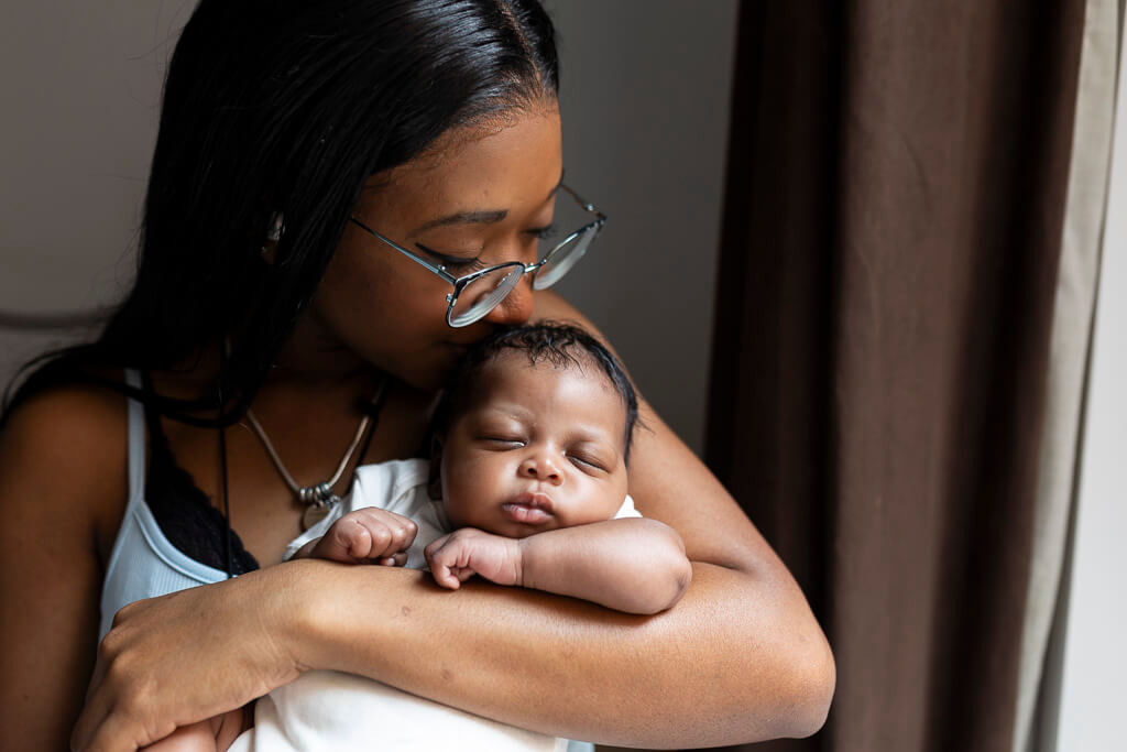 Mother kissing her sleeping newborn baby while holding her in her arms during a newborn photoshoot at home in Glasgow