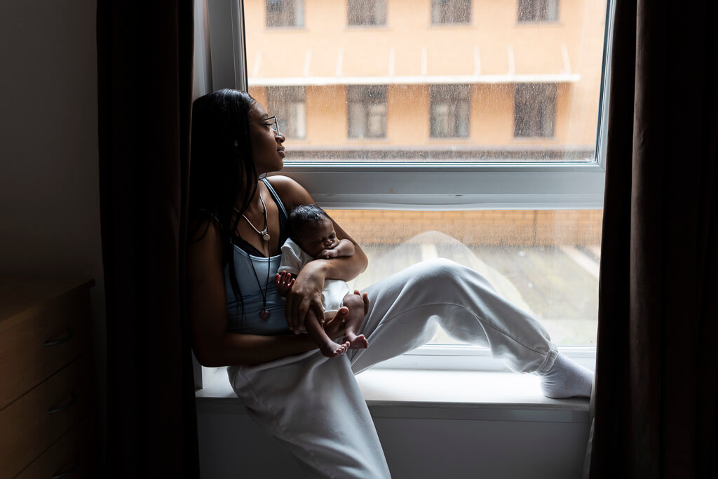 Mum sitting beside a window, gently holding her newborn baby in natural daylight