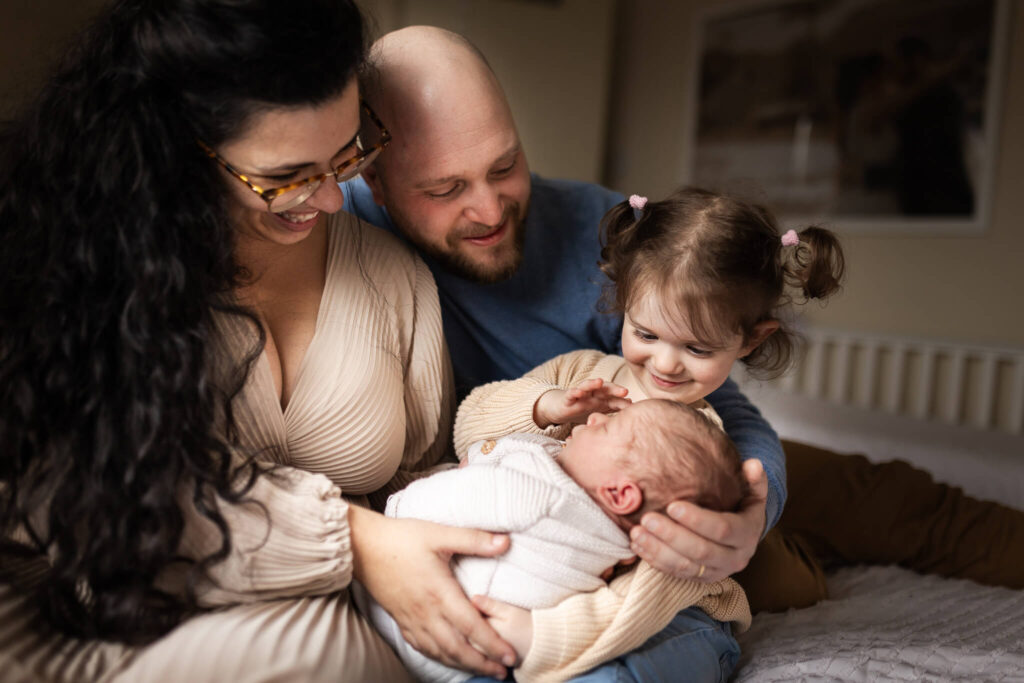 Family of four sitting together as older sister gently strokes their newborn baby during a family newborn photoshoot in Glasgow