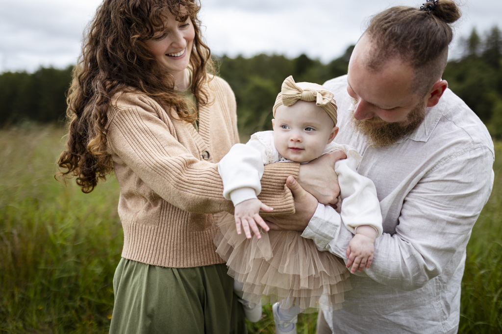 Parents with their toddler posing on a meadow during an outdoor family photoshoot in Glasgow, wearing coordinated beige and white outfits with layered knitwear