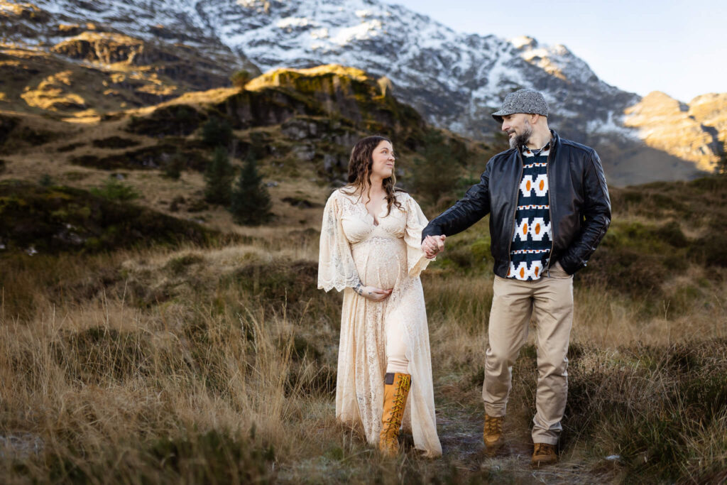 Couple walking hand in hand and looking at each other during a maternity photoshoot in Glasgow, with Scottish mountains in the background