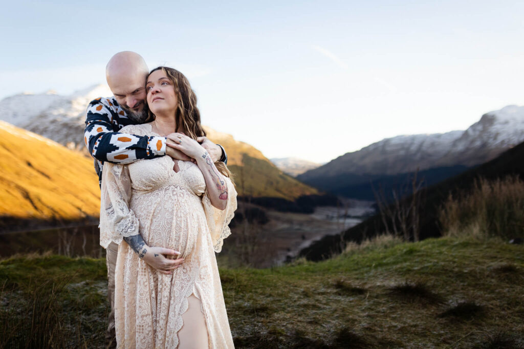 Couple embracing against a mountain landscape during a maternity photoshoot in Scotland, with the woman wearing a lace dress