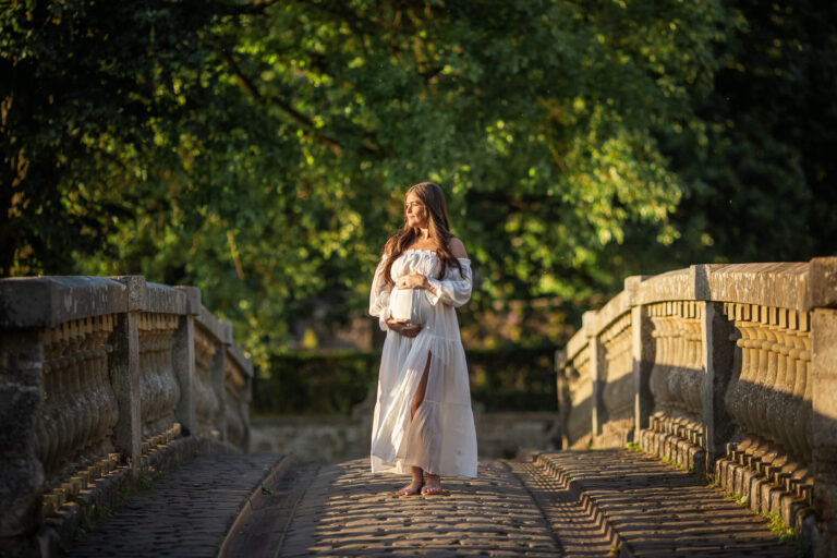 Pregnant woman in a white dress standing on a stone bridge during an outdoor maternity photoshoot in Glasgow