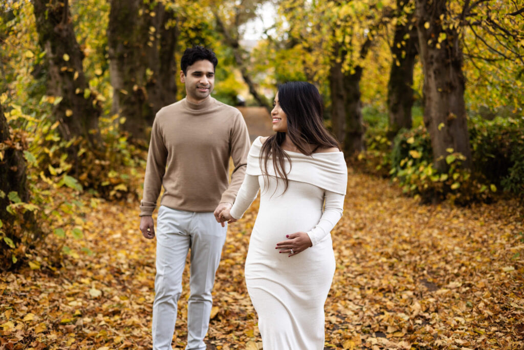 Couple walking hand in hand through an autumn park during a maternity photoshoot in Glasgow, surrounded by yellow leaves