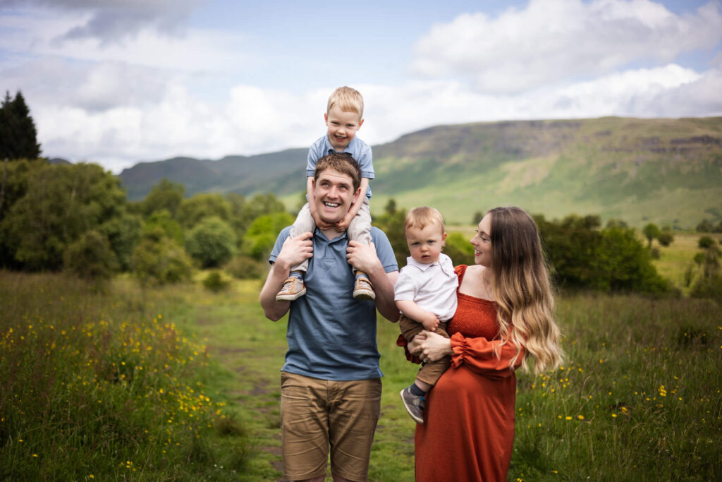 Family of four walking across a meadow, with one child riding on his father’s back.