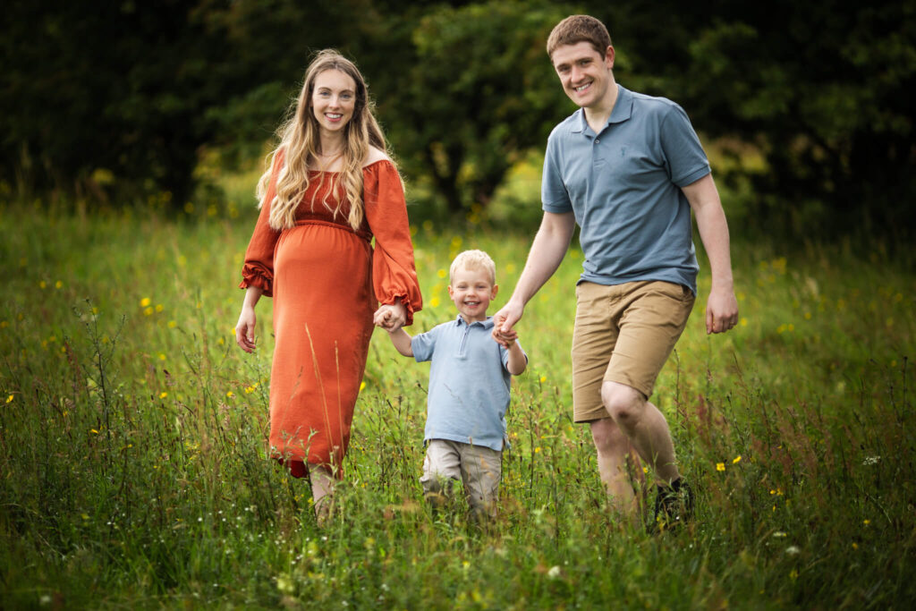 Expectant parents holding their young son's hands as they walk through a meadow during a family maternity photoshoot in Glasgow