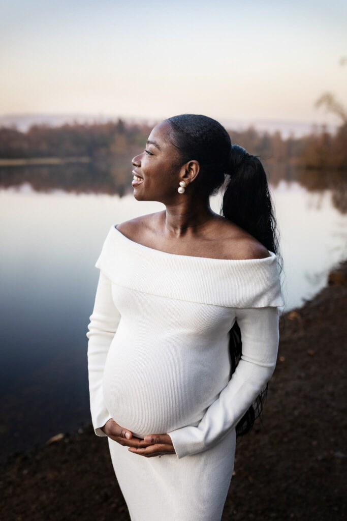 Pregnant woman in a white off-shoulder dress by the water at sunset during a maternity photoshoot in Glasgow
