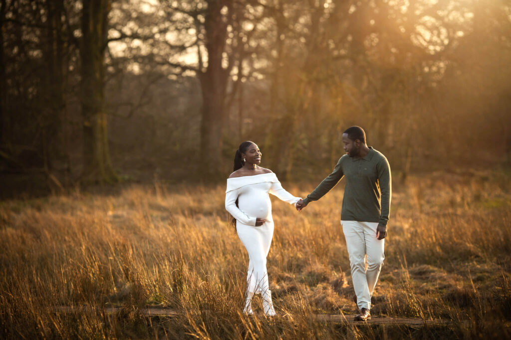 Expectant parents walking through golden grass at sunset in Mugdock Country Park during a natural outdoor maternity photoshoot.