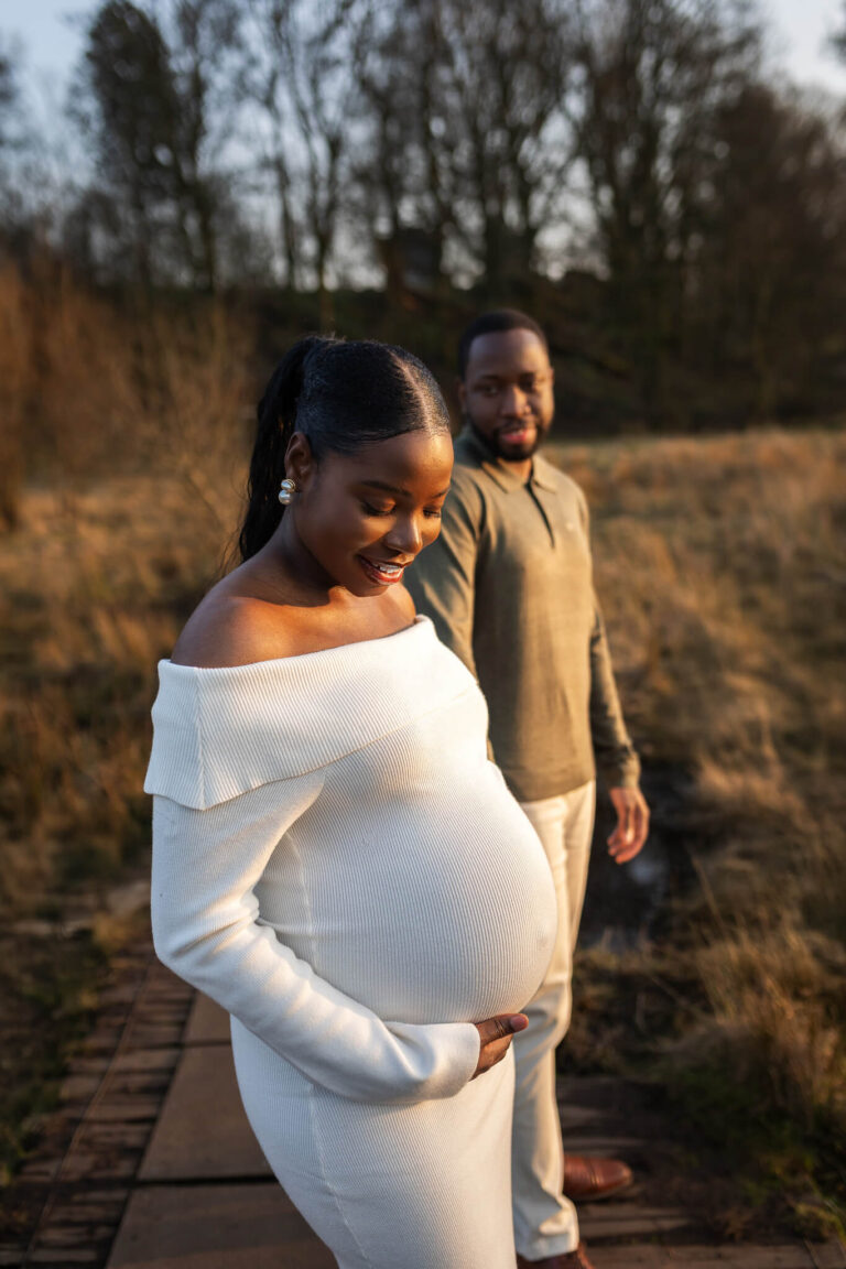 Pregnant woman in a white knit dress smiling down at her bump beside her partner during an outdoor maternity photoshoot in Glasgow