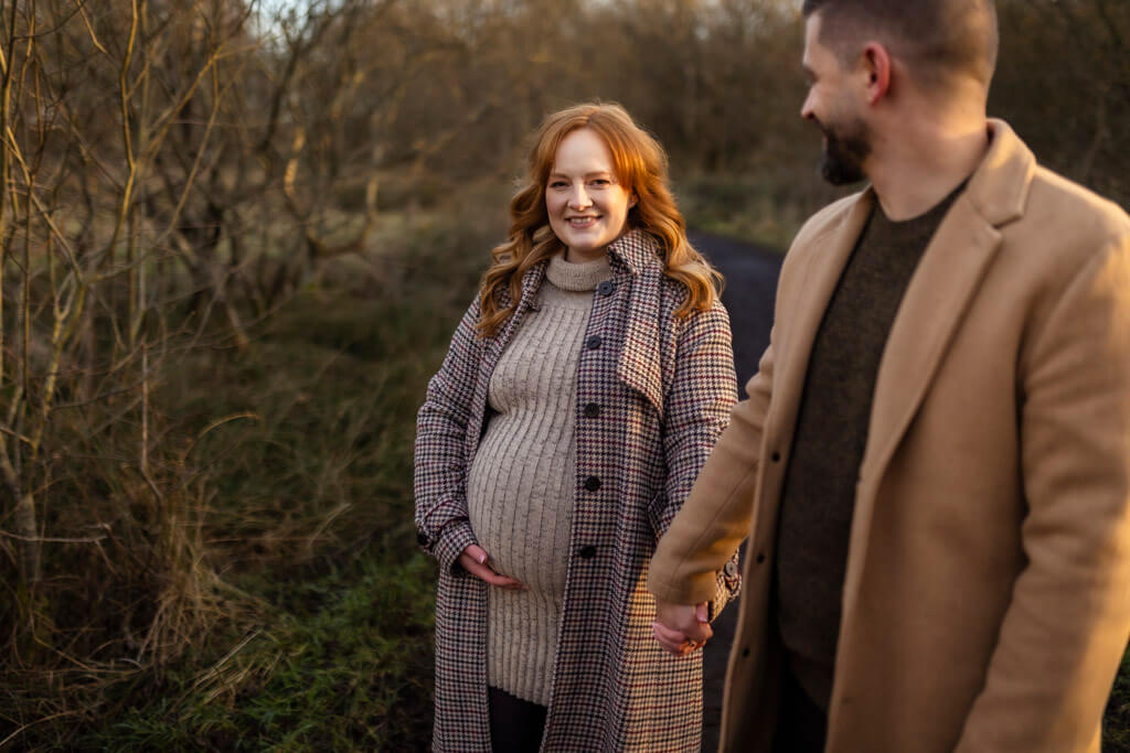 Pregnant woman smiling and holding her partner’s hand during a winter maternity photoshoot in Glasgow, wearing a winter coat and textured grey dress