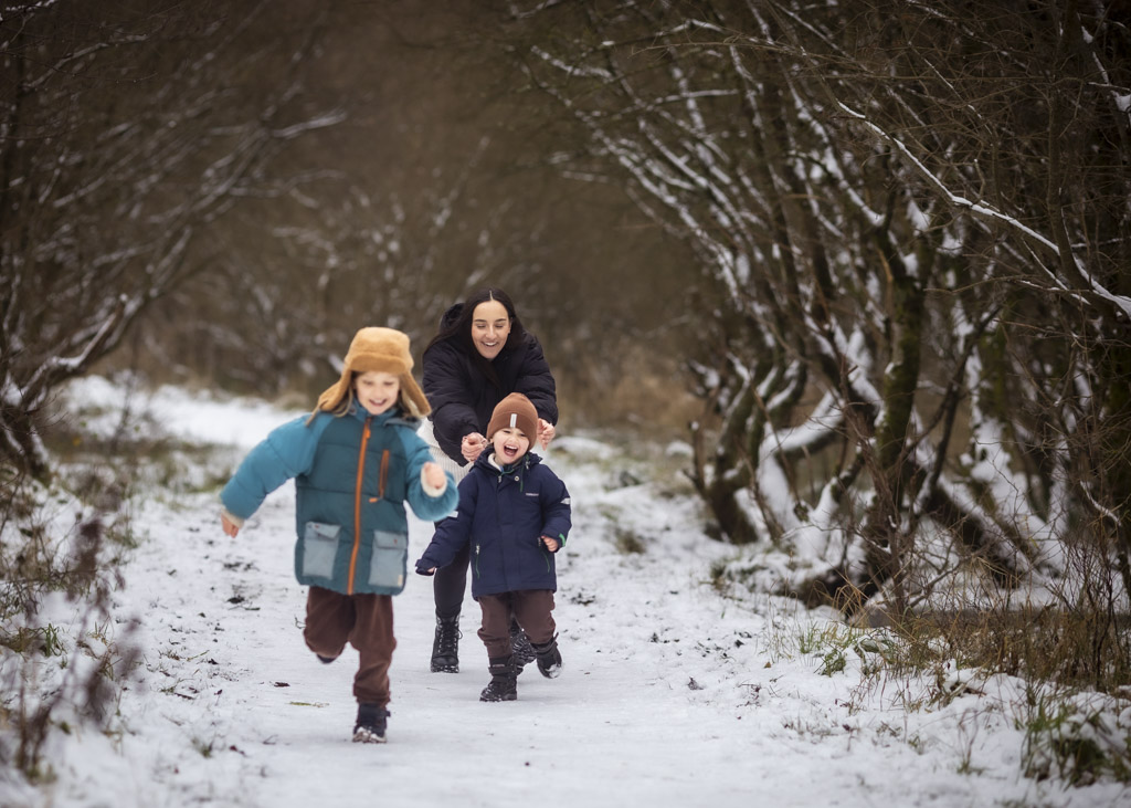 Mother running with her two children along a snowy woodland path during a winter family photoshoot in Glasgow