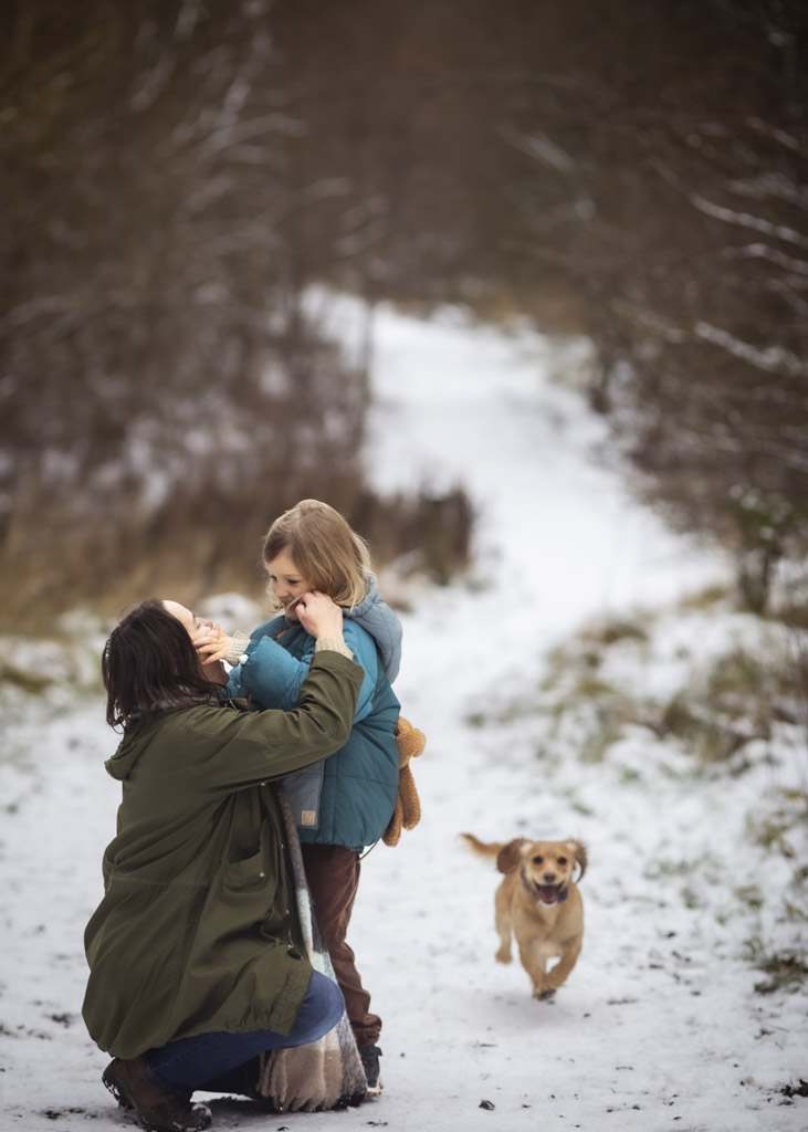 Mother strokes her face and hugs her daughter during a snowy winter family photoshoot in Glasgow, with their dog nearby
