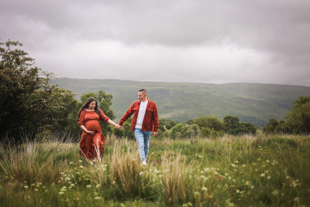 Pregnant woman in an orange dress walking hand in hand with her partner across a hillside in the Scottish countryside during a maternity photoshoot