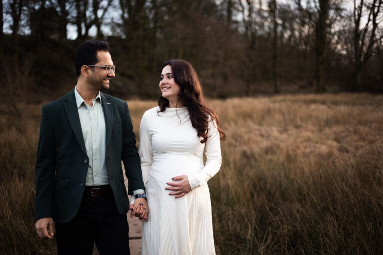 Pregnant woman in a white dress holding her bump while looking at her partner in a meadow during an outdoor maternity photoshoot in Glasgow