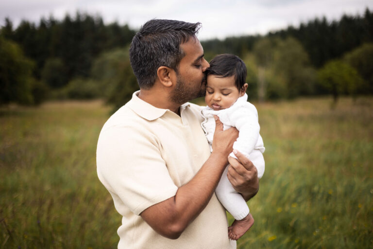 Father kissing his newborn baby while holding him in a meadow during a summer outdoor newborn photoshoot in Glasgow