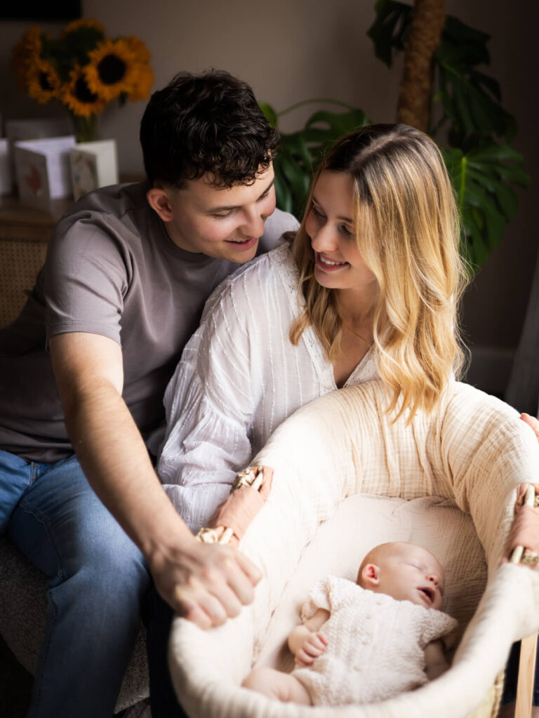 New parents smiling at each other on the sofa while their newborn baby rests in a Moses basket