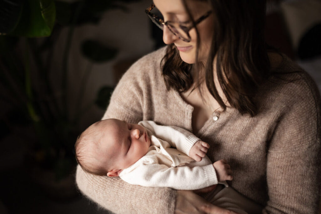 Mum in glasses cradling her sleeping newborn baby in a softly lit room at home