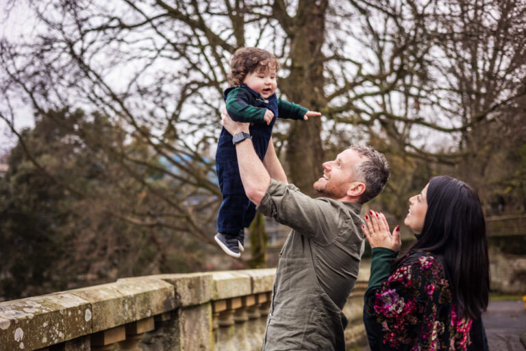 Father lifting his young son as mum watches during an outdoor family photoshoot in Glasgow